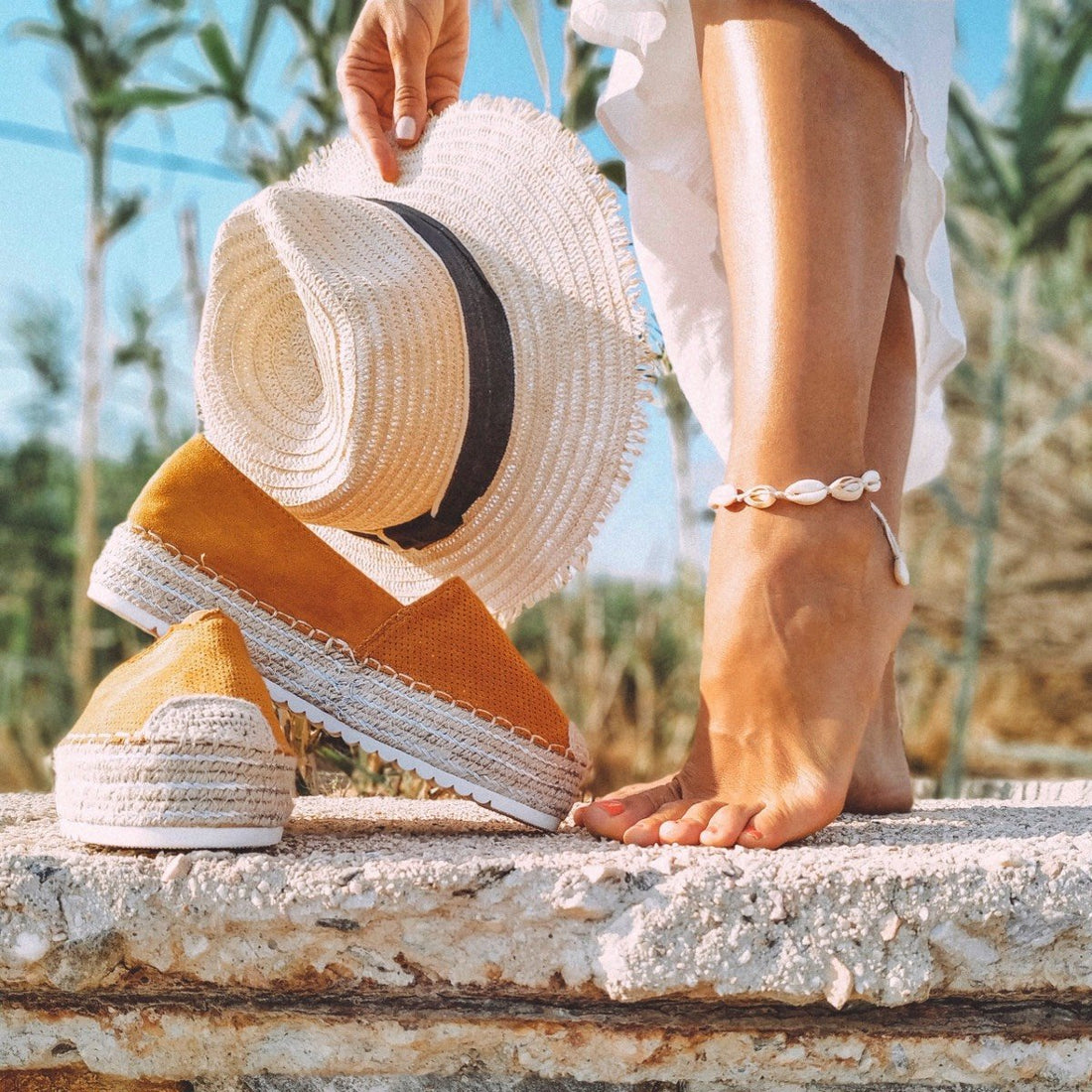 The feet of a barefoot women wearing a seashell ankle bracelet an orange pair of espadrilles are visible and a straw hat with a black band is in her hand